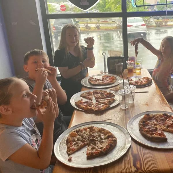 Four children happily eating Green Fire Pizza at a table by a window on a rainy day.
