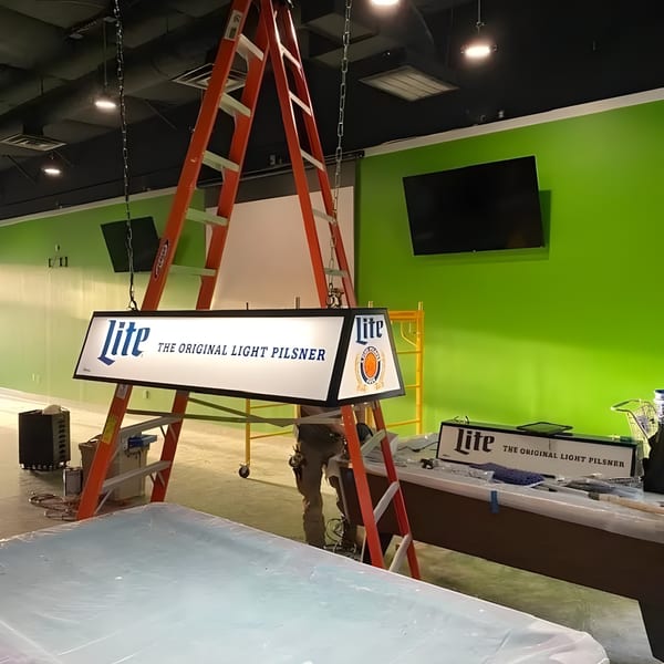 A Lite beer sign hangs from the ceiling, with a person on a ladder working on it.