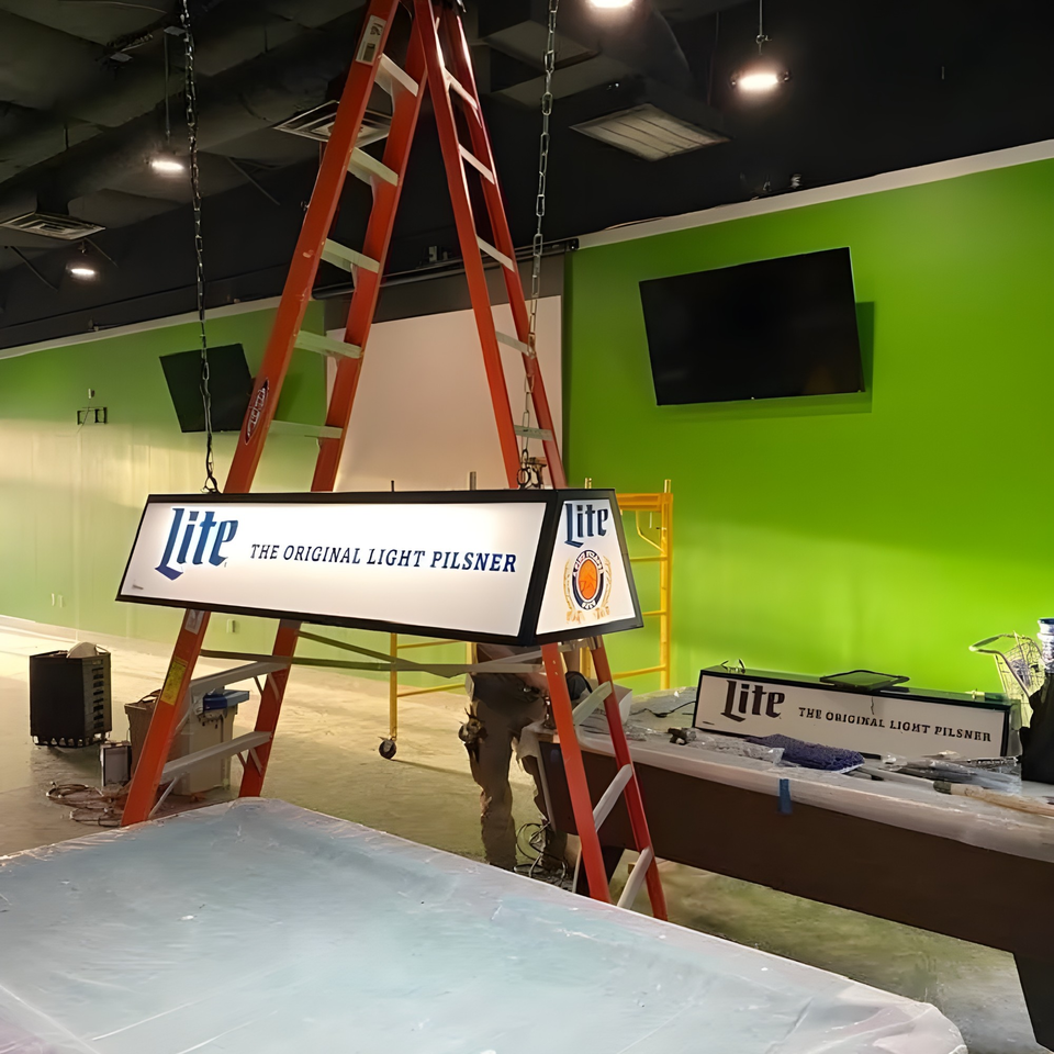 A Lite beer sign hangs from the ceiling, with a person on a ladder working on it.