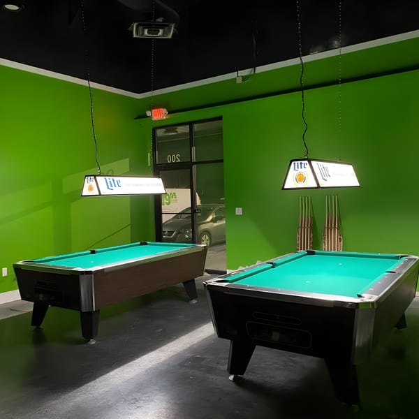 Two pool tables with green felt and Miller Lite lamps hang above in a room with green walls.